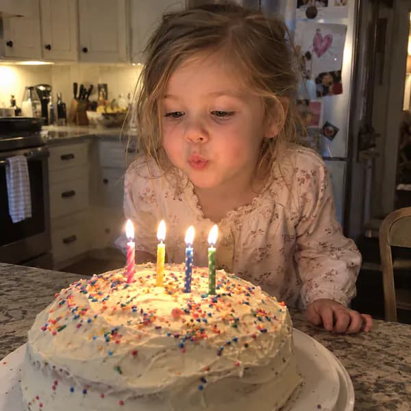 AI-generated photo: a little girl blows out four candles on a homemade sprinkle cake in a kitchen.